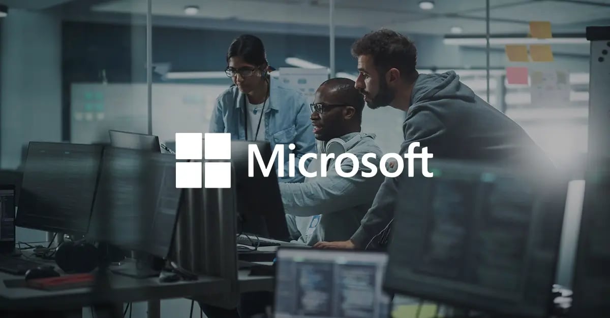 Photo of three people sat or standing around a computer screen in an office with a white version of the Microsoft logo superimposed in the middle foreground